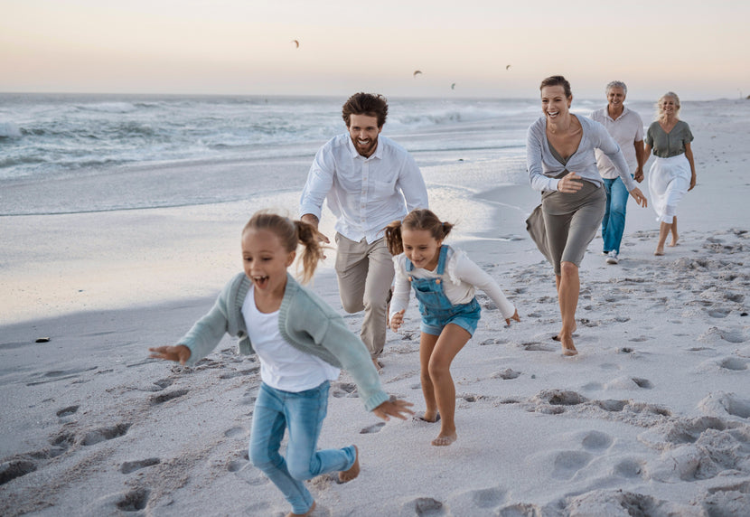 Famille multigénérationnelle courant et s’amusant ensemble sur une plage au coucher du soleil, avec deux jeunes filles en tête, suivies par leurs parents et grands-parents souriants.